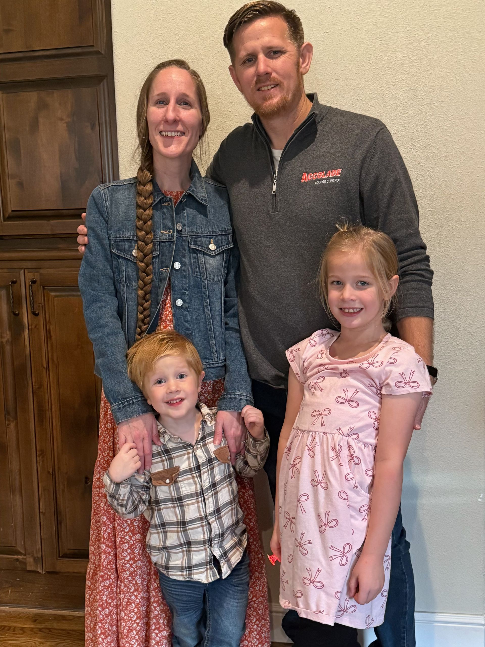 A family of four smiling in an indoor setting with wooden cabinets in the background.