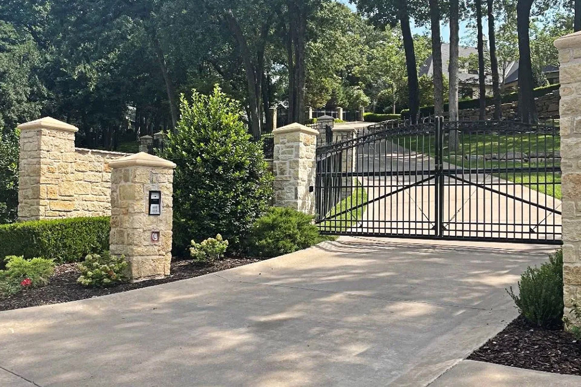 Stone entrance gate with black iron bars, leading to a driveway, set in a tree-lined area.