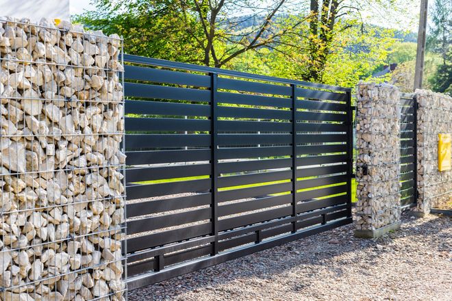 Black horizontal slat gate with stone pillars in a gravel driveway.