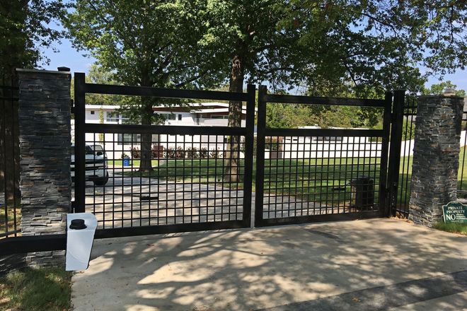 Black metal gate with stone pillars opens onto a driveway and a white house with trees in the background.