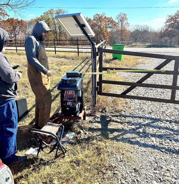 Two people installing a solar-powered gate opener at a wooden gate. A person is holding a long bar. Sunny outdoor setting.
