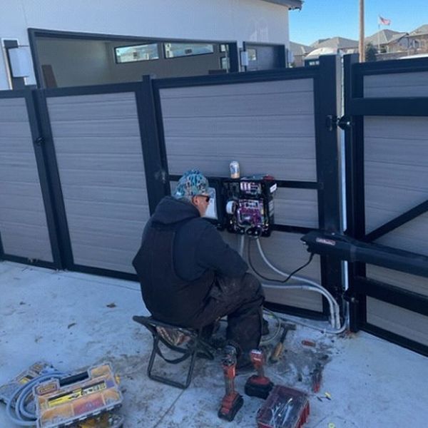 Man working on gate opener, sitting on a stool. Tools and materials on concrete. Gray and black gate.
