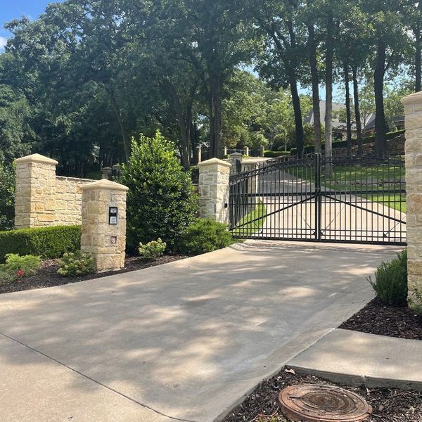 Stone entrance with black gate, pillars, and a concrete driveway leading to a house, trees in background.