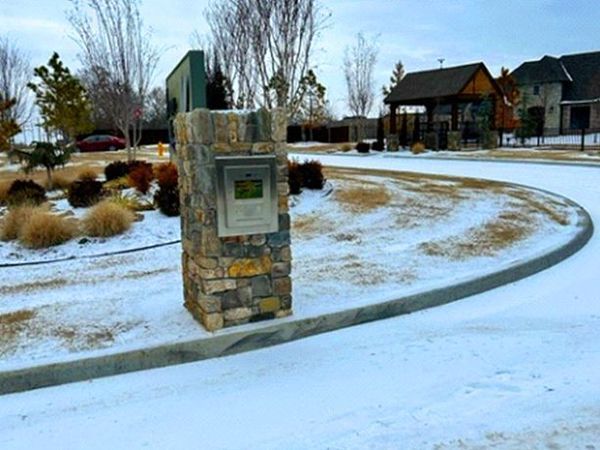 Stone pillar with electronic panel in snow. Curved driveway, houses and trees in the background.