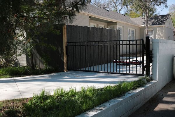 Black metal gate leading into a concrete driveway, bordered by a white wall and greenery, house in background.