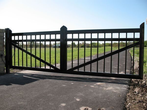 Black metal driveway gate on paved road, with a field in the background.