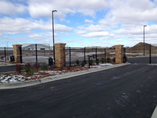 Gated entrance with black metal fence and tan brick pillars, with a cloudy sky backdrop.