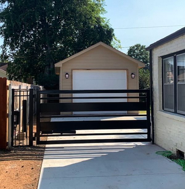 Black gate in front of a tan garage with a white door. Concrete driveway.