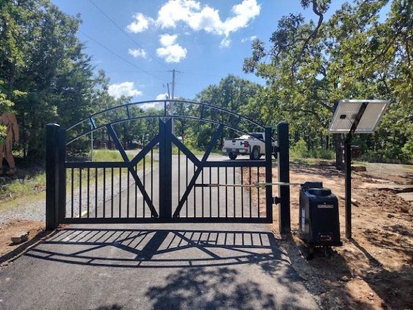 Black metal gate at a driveway entrance, with solar panel and a white truck visible beyond.