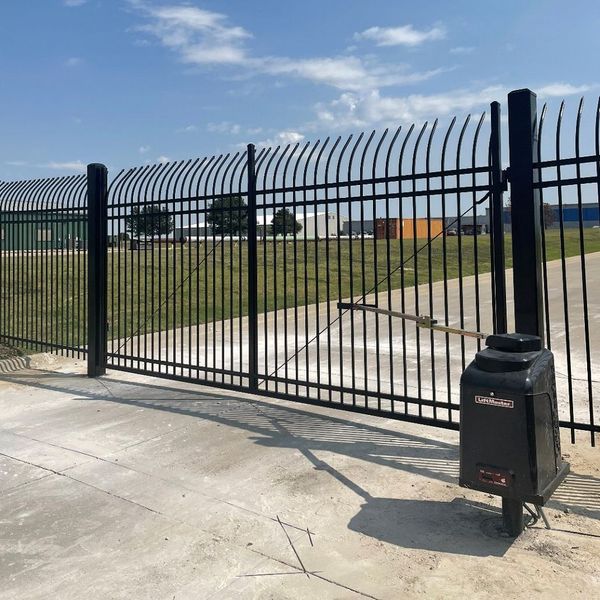 Black security gate with a curved top, set on a concrete surface. An automatic gate opener is attached.