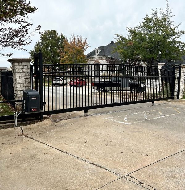 Black metal gate, automatic opener, brick pillars, driveway, cars, and trees in background.
