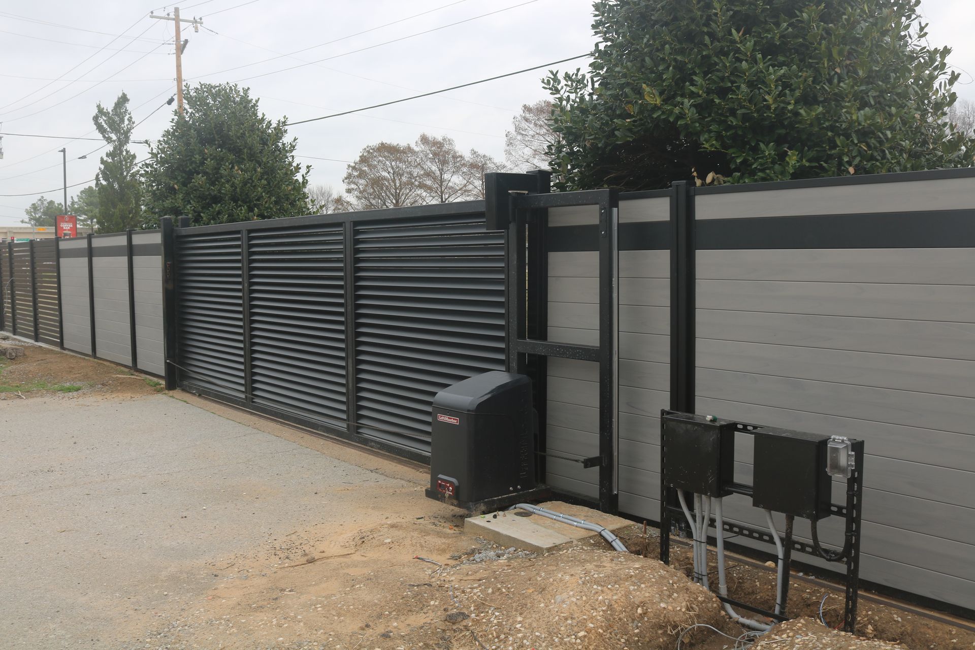 A sliding gate with dark metal slats and gray privacy panels, featuring a motor and control boxes at an outdoor entrance.