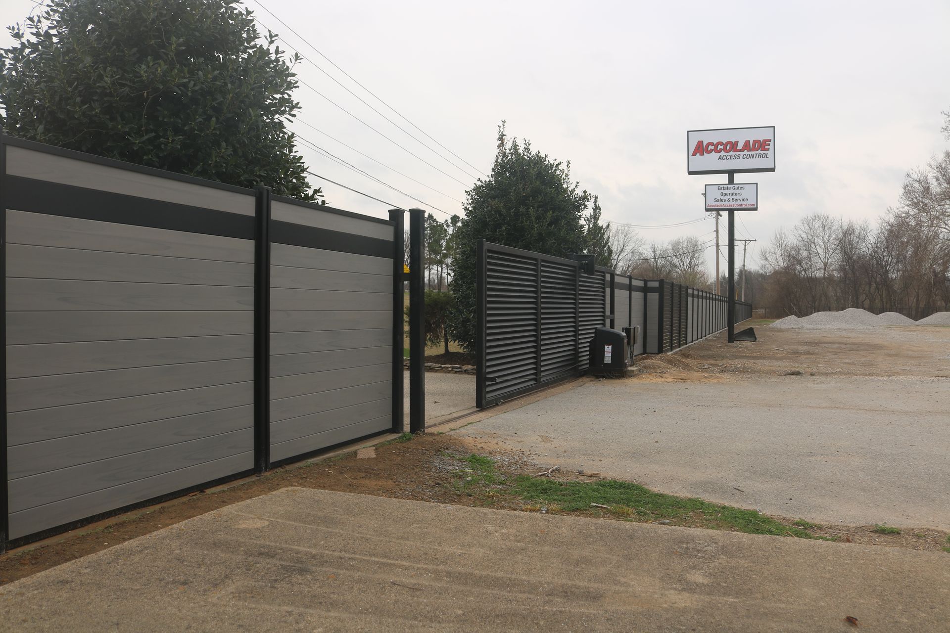 Modern gray and black panel fencing stands beside a gated entrance with an Accolade signage post on a gravel lot.