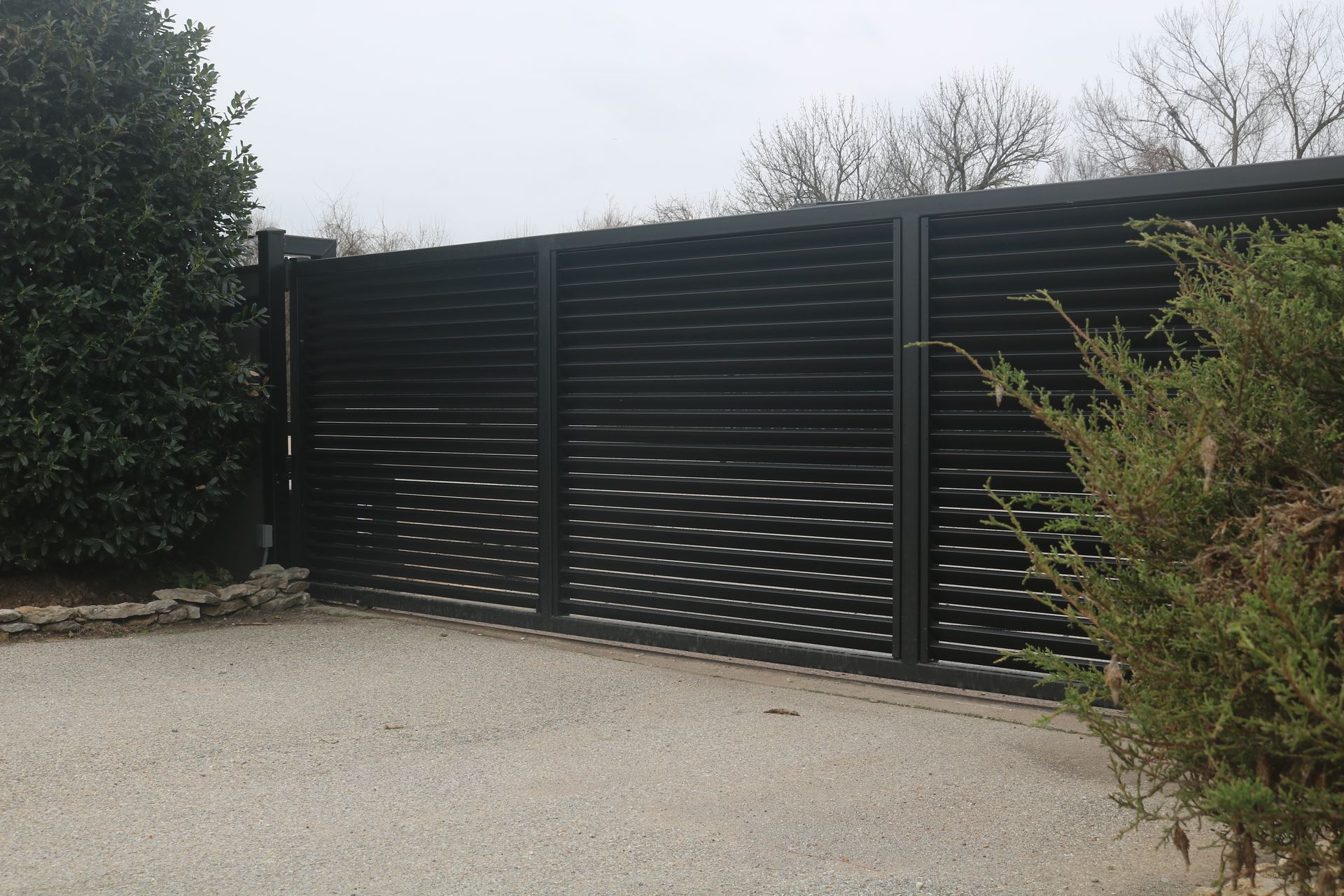 A modern black metal gate with horizontal slats installed in a gravel driveway.