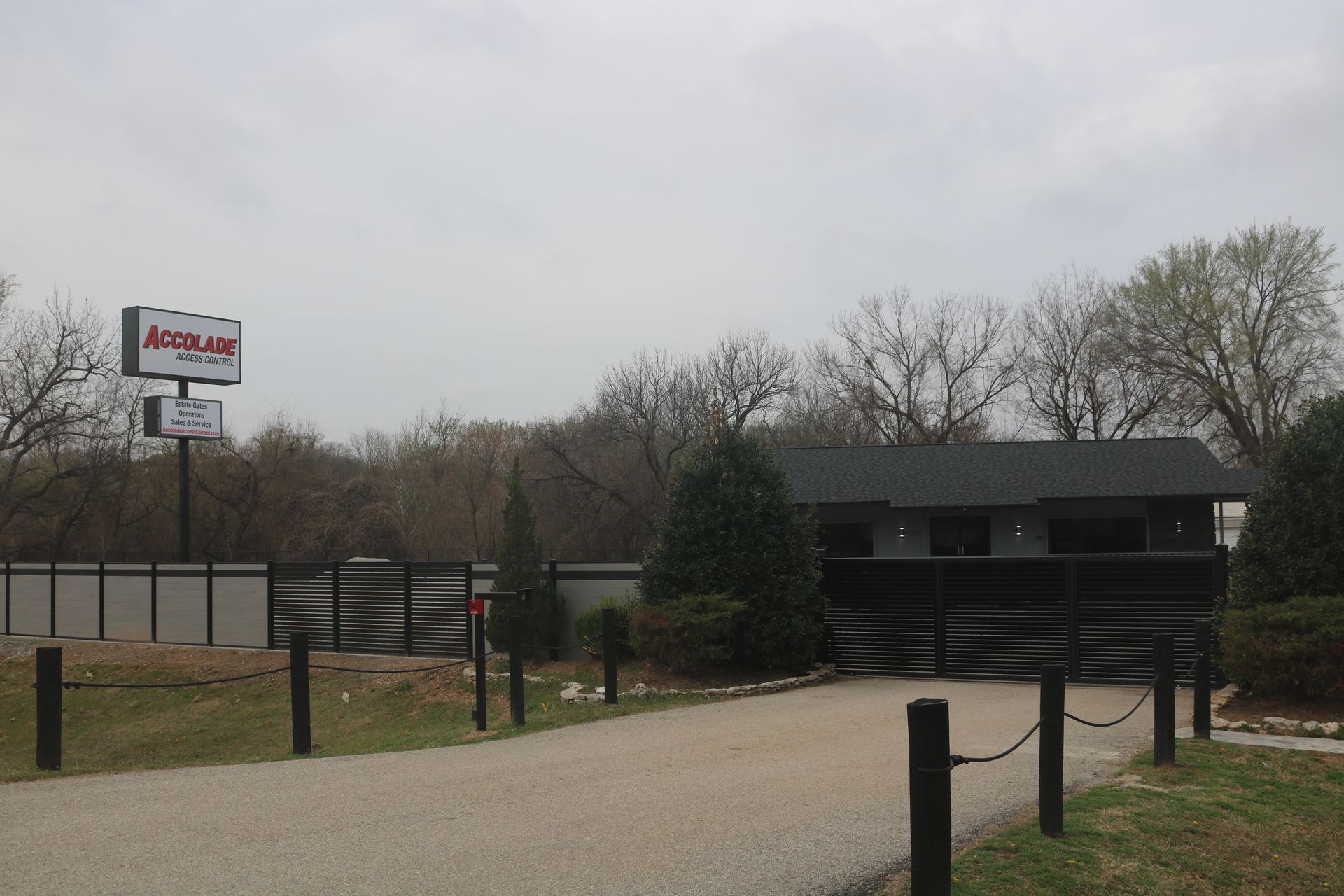 An Accu-Lab sign stands above a paved entrance drive leading to a dark building behind a black metal fence and landscape.