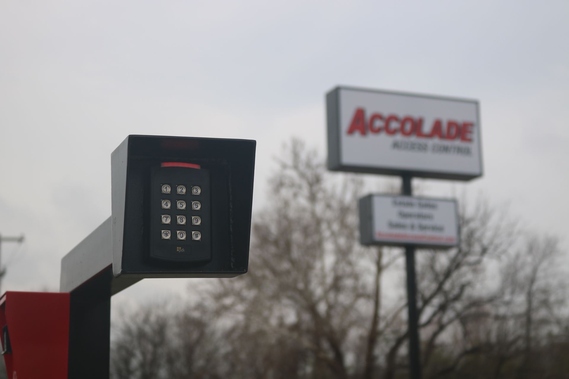 A keypad mounted on a pedestal for an Accolade access control gate, with a blurred sign in the background.