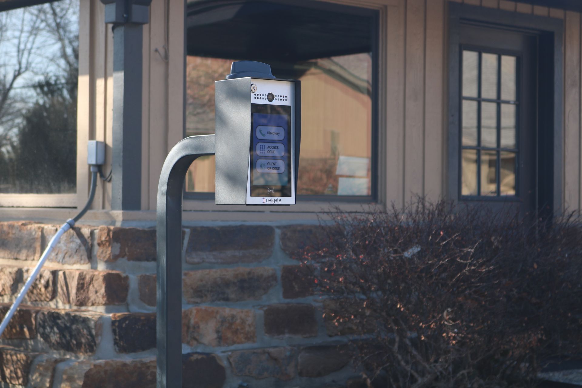 A digital gate intercom system on a metal post in front of a building with a stone foundation and a glass door.