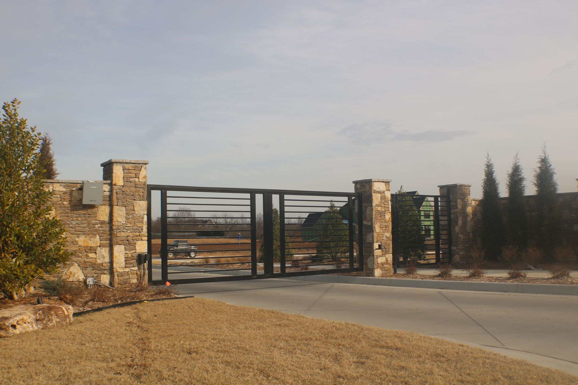 Modern black metal double gates between stone pillars, leading to a flat, open outdoor lot under a bright, hazy sky.