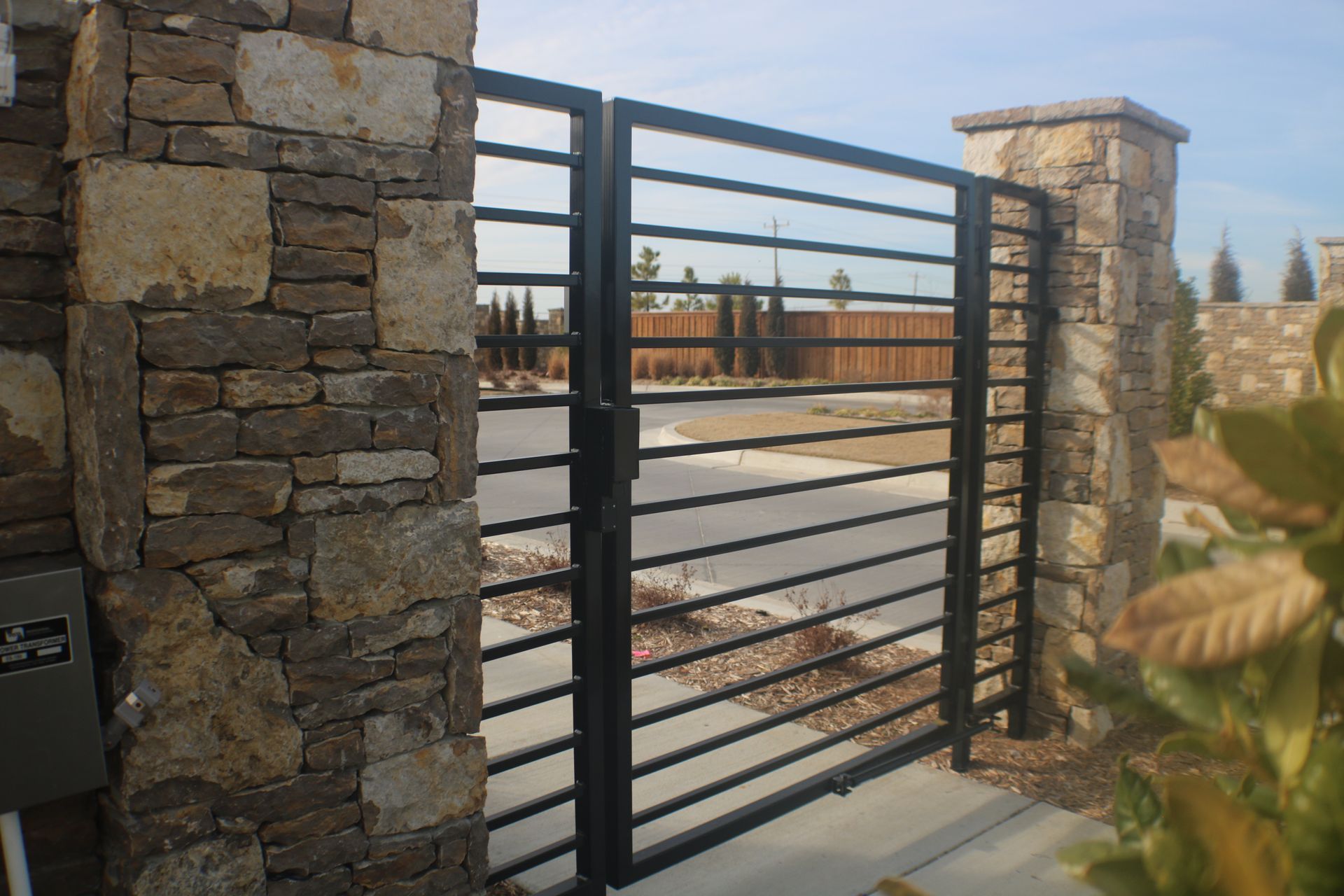 A black horizontal metal gate secured between two rustic stone pillars on a concrete driveway.