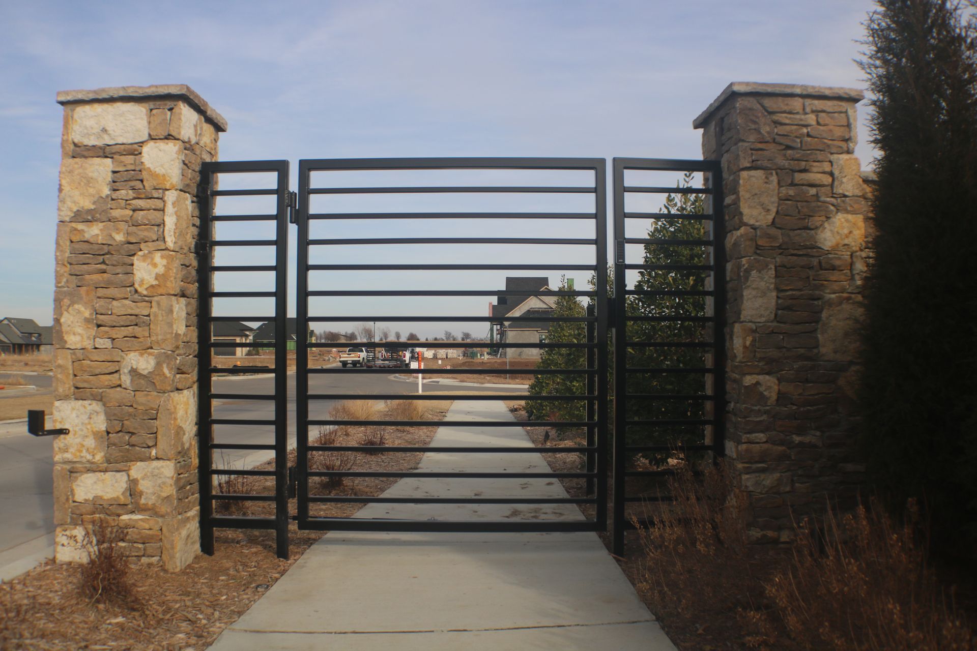 Black metal pedestrian gate between two stone pillars along a concrete walkway.