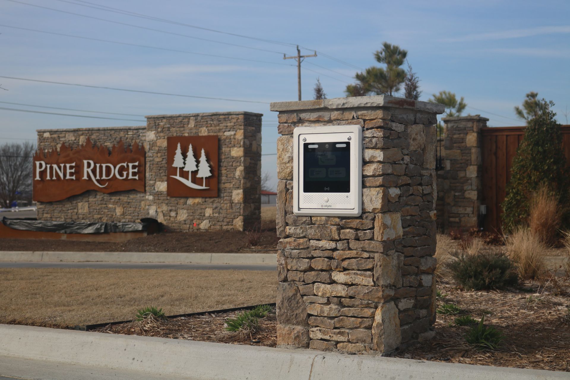A stone entrance sign for Pine Ridge with a pine tree logo, situated outdoors near a road under a blue sky.
