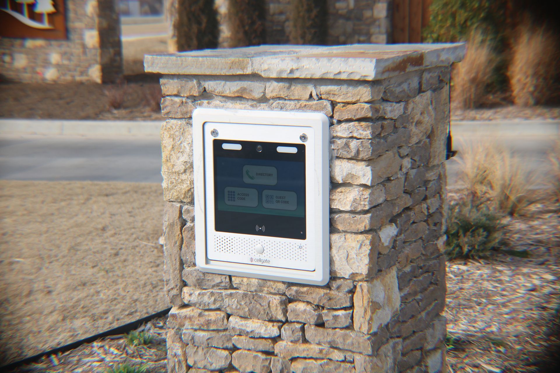 A stone pillar entrance gate intercom system featuring a white rectangular interface with a screen and a speaker.