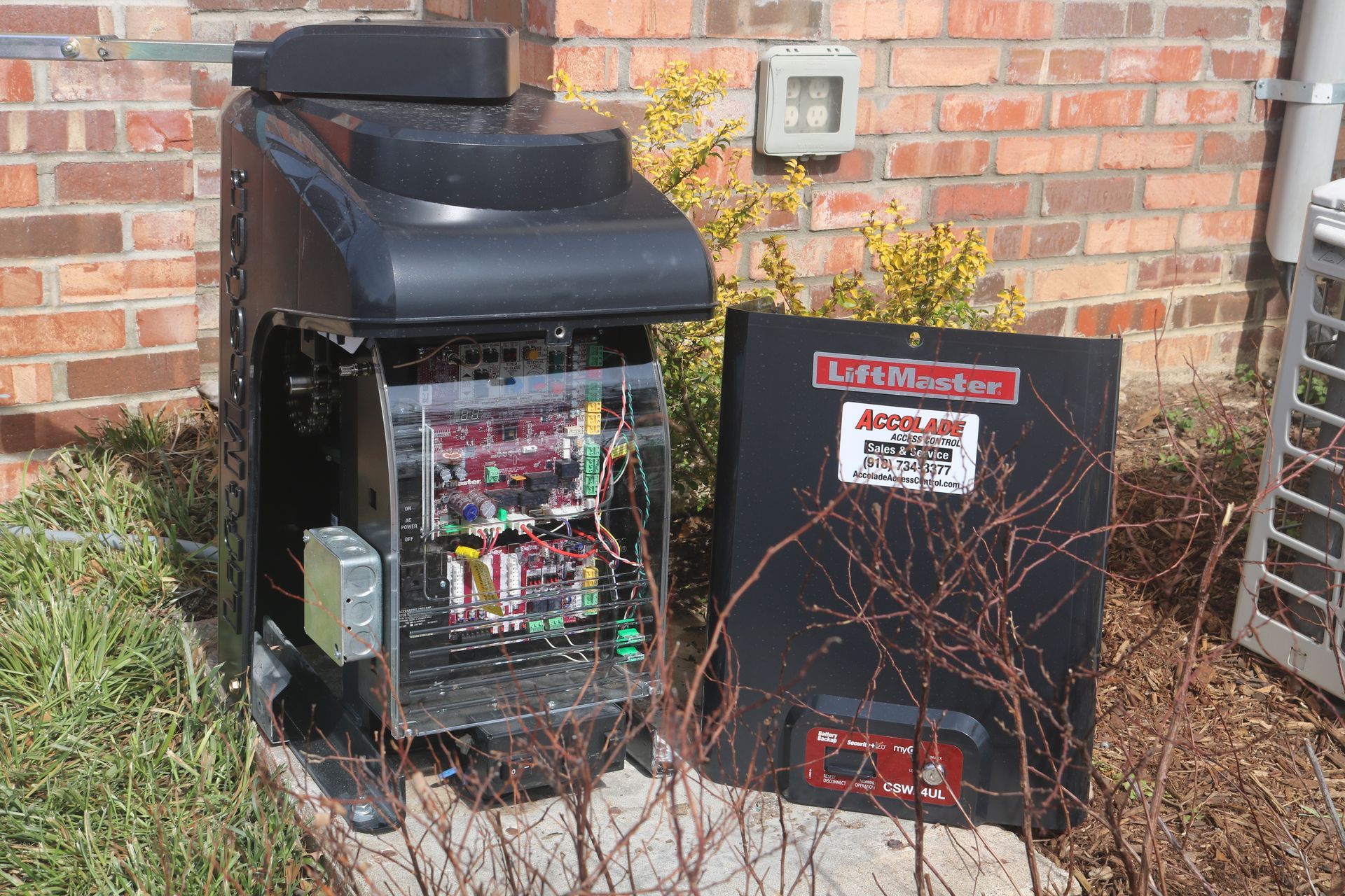 A black LiftMaster gate operator unit with its cover removed, revealing internal circuit boards, sitting on a concrete pad.