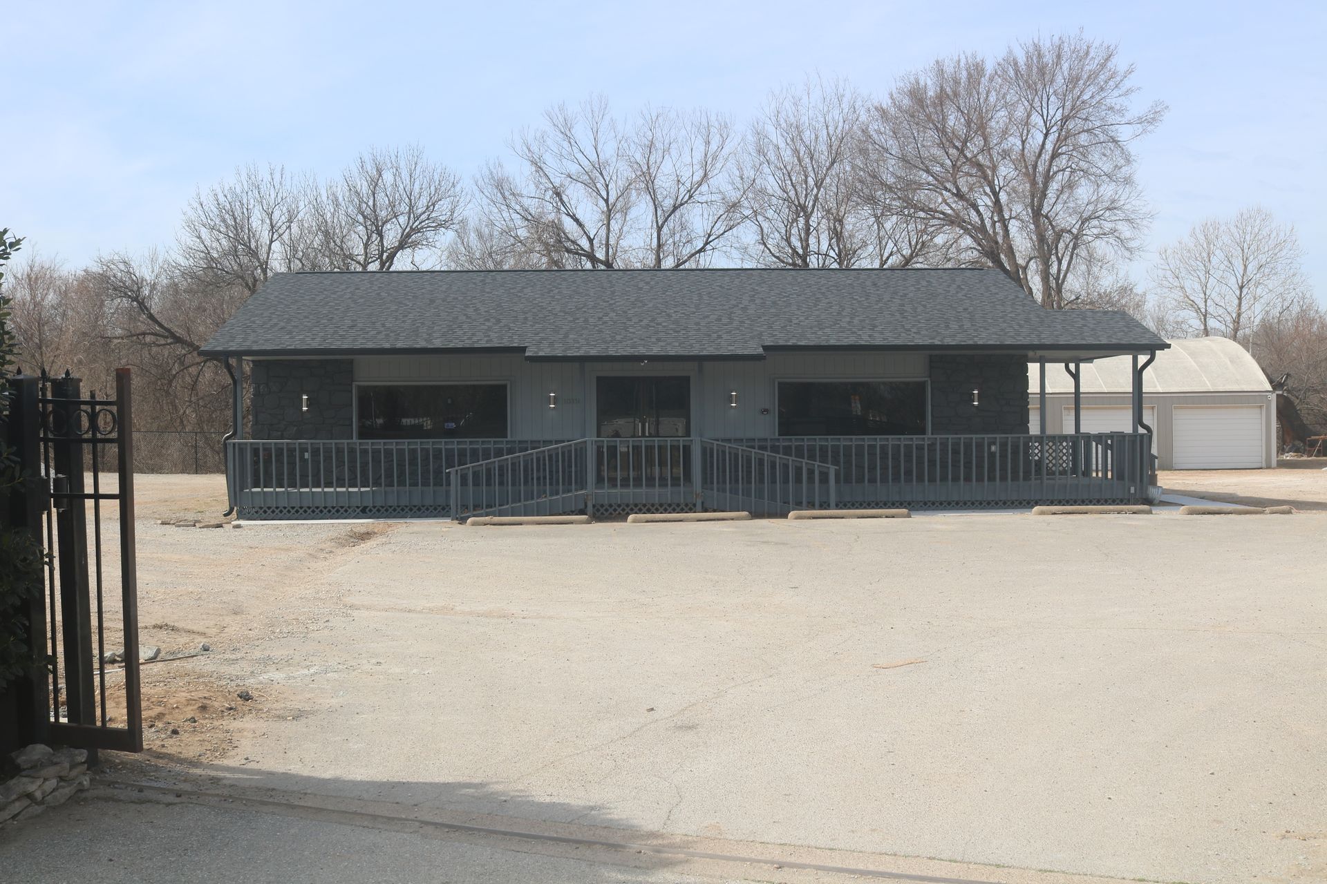 A gray single-story commercial building with a wide front deck and dark roof sits on a gravel lot under a clear sky.