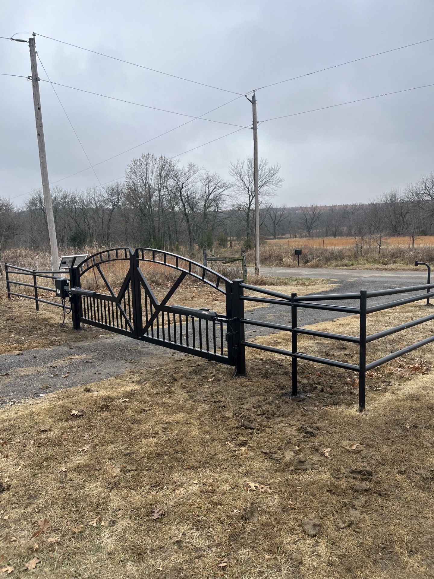 A black metal arched driveway gate with matching three-rail fencing, situated in a rural, grassy field under a grey sky.
