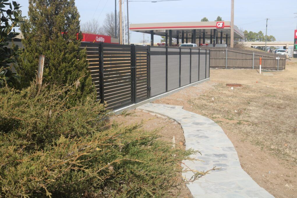 A modern, black-slatted privacy fence runs alongside a curved, light-colored concrete path near a gas station.