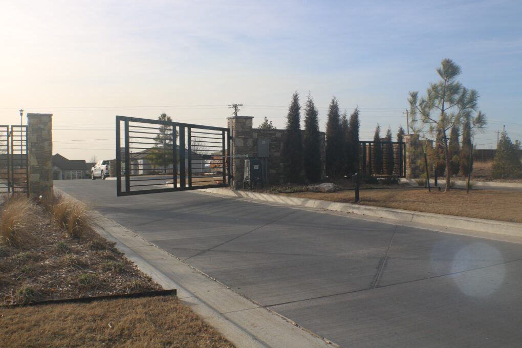 A wide metal gate stands open at the entrance to a paved driveway, flanked by stone pillars and pine trees.