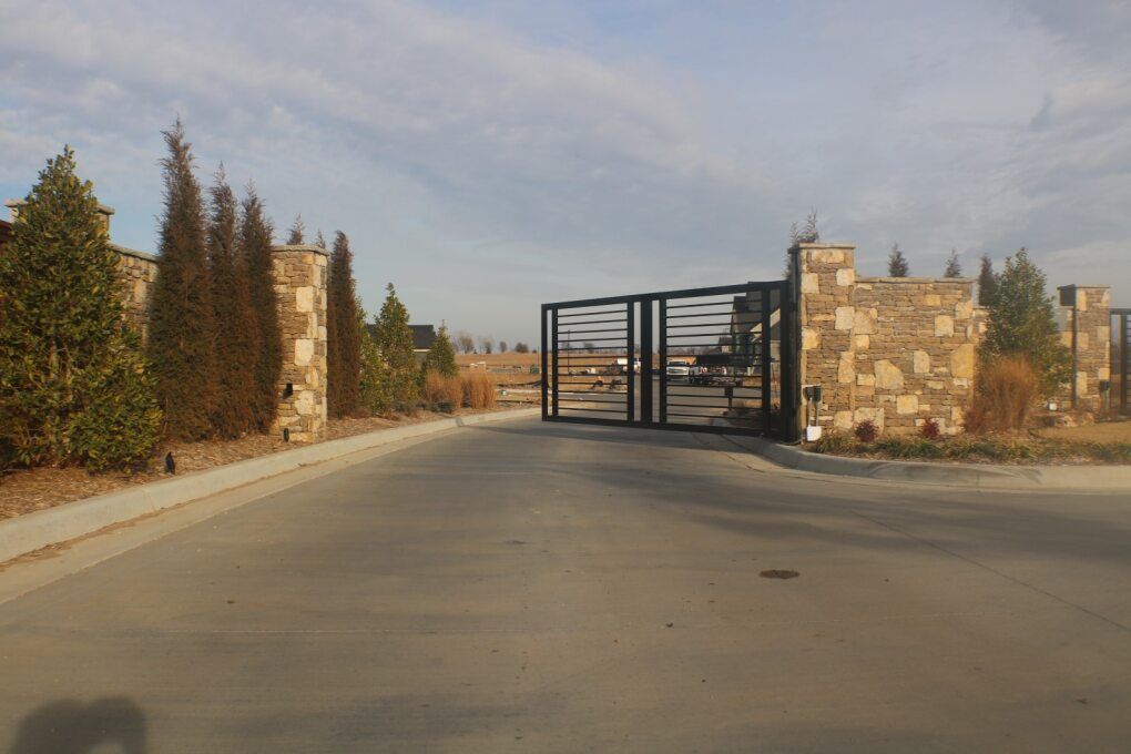 A modern, open black metal gate sits between two stone pillars on a paved road, flanked by evergreen trees.