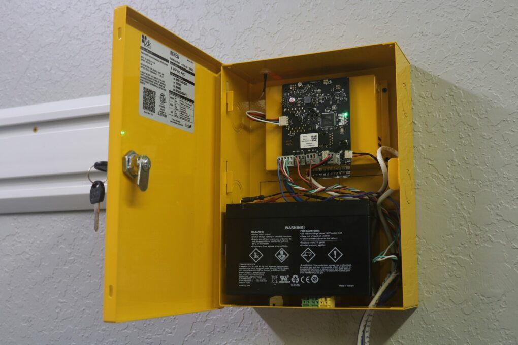 A yellow wall-mounted cabinet with the door open, showing an internal electronic control board and a black battery backup.