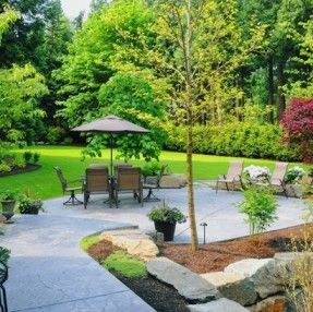 Patio with umbrella, table, and chairs in a lush garden with trees and green lawn.
