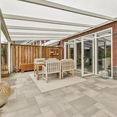Outdoor patio with seating under a white-framed roof, adjacent to a red brick building with glass doors.