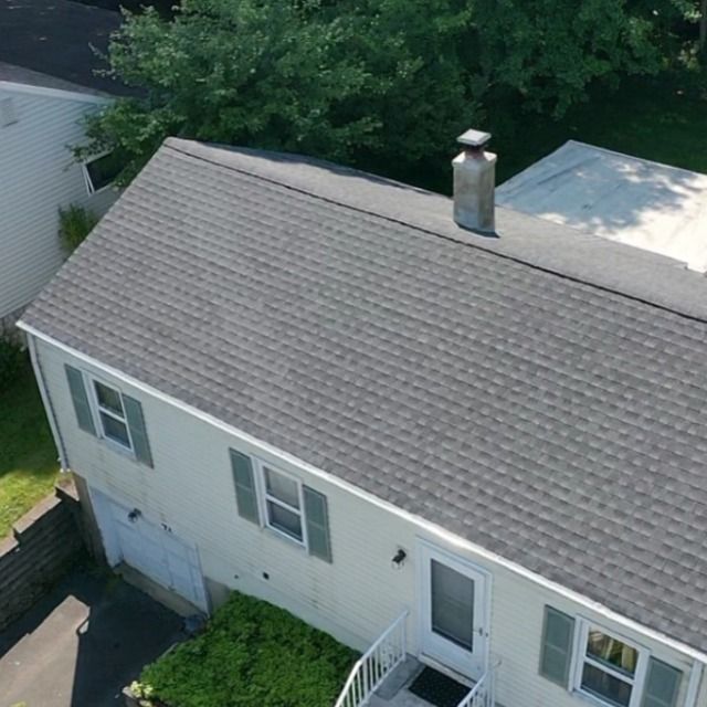 Overhead view of a two-story house with a gray shingled roof, light siding, and green shutters. A chimney rises from the roof.