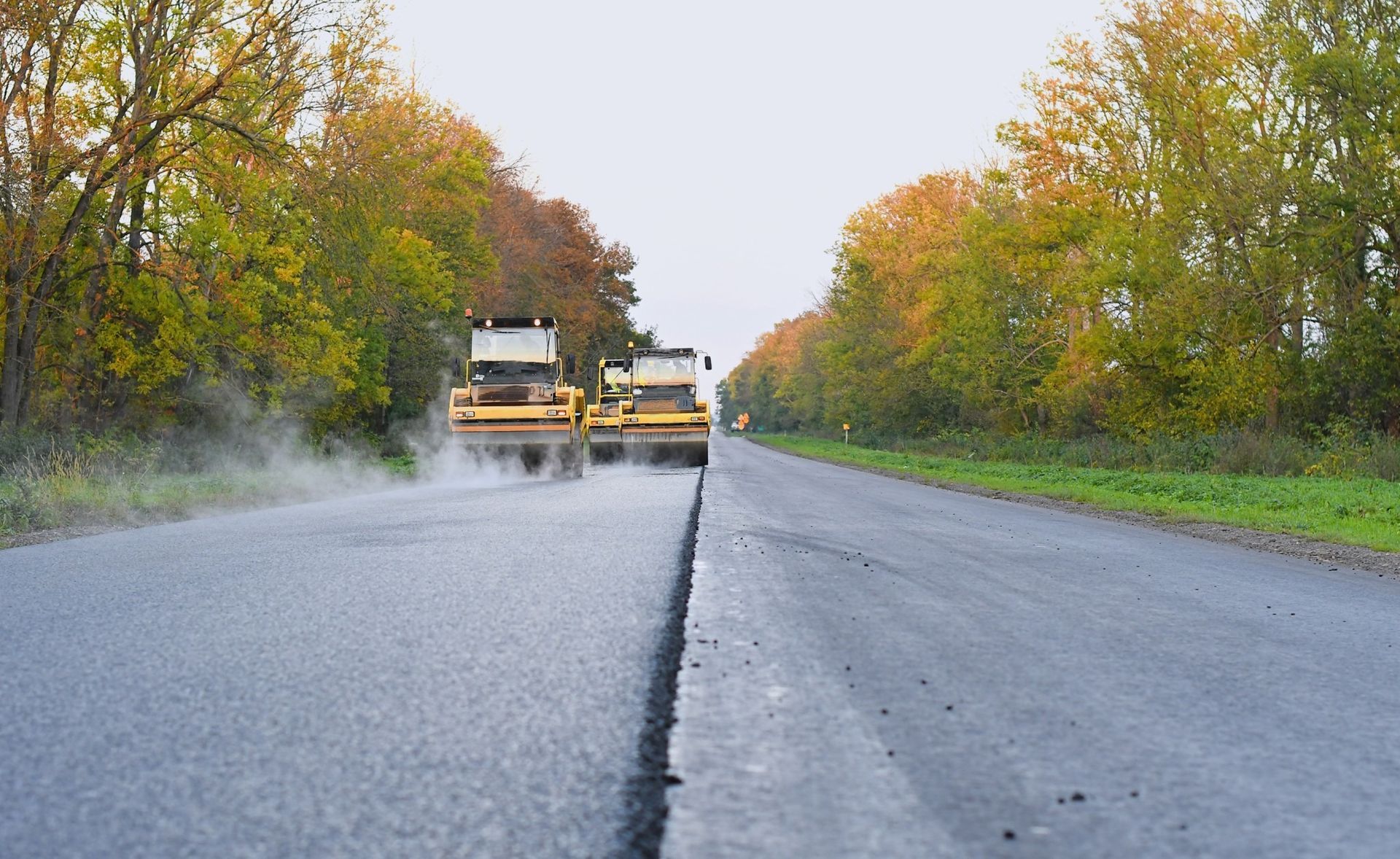 Road maintenance trucks spraying asphalt on a road with a central crack, lined by trees.