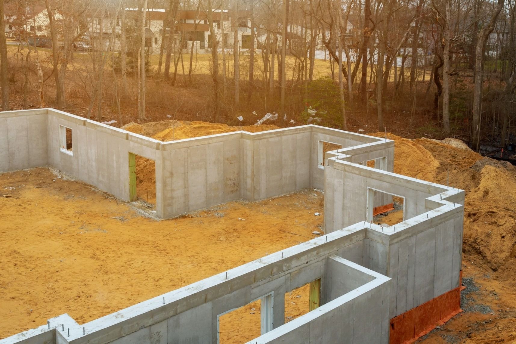 Concrete foundation walls of a house under construction, in a dirt lot surrounded by trees.