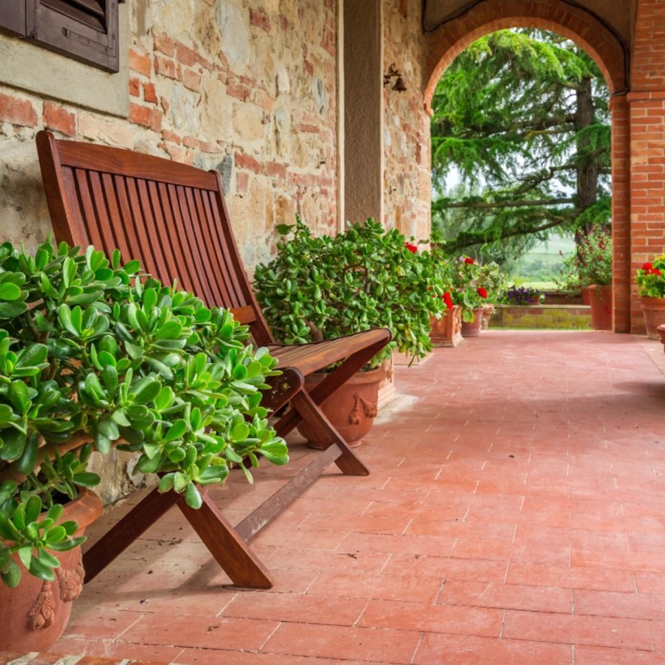 Wooden chair on a terracotta-tiled porch with potted plants and a brick and stone wall. An arched doorway offers a view of trees.