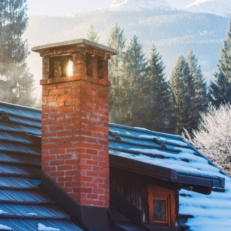 Brick chimney on a snow-covered roof against a backdrop of snow-dusted evergreen trees and mountains.