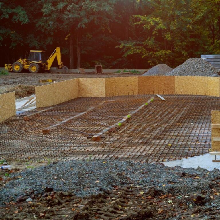 Construction site with a yellow backhoe in the background, wooden forms outlining a foundation, and a metal rebar grid laid down, ready for concrete.