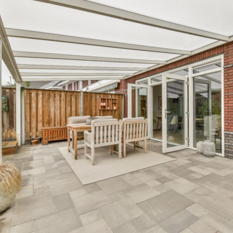 Patio with seating under a white canopy, adjacent to a brick house with glass doors. Wooden fence in the background.