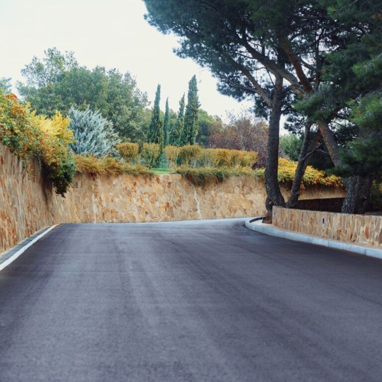 Asphalt road curving uphill, bordered by a stone wall and landscaping with trees.