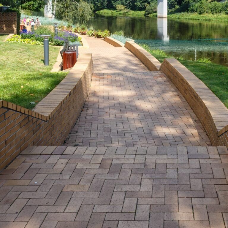 Brick pathway leading down to a river, with brick walls on either side. Lush greenery and flowers flank the path.