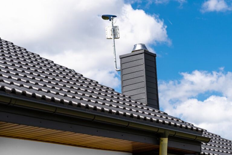 Black roof tiles with dark gray chimney and antenna against a partly cloudy sky.