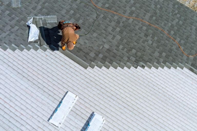 Aerial view of roofers installing shingles on a light gray roof. One worker in a brown coat is kneeling while the other is nearby.