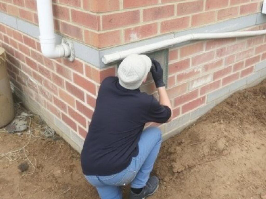 Person in black shirt and jeans working on brick building exterior, near ground level, wearing gloves.