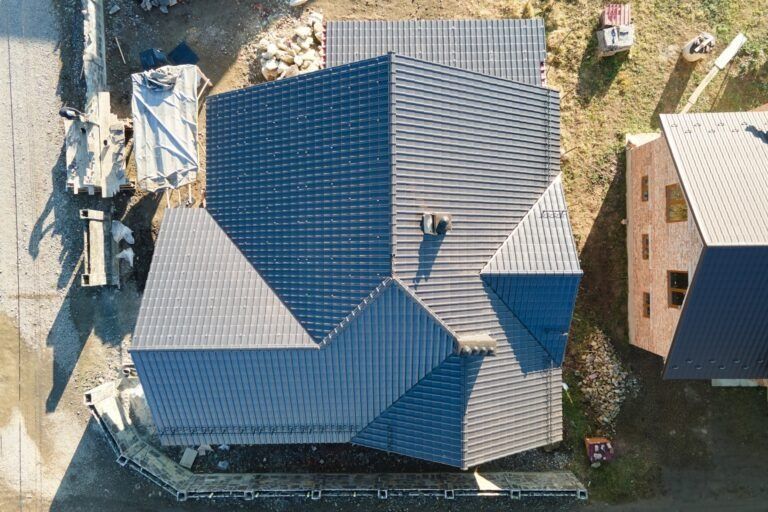 Overhead view of a house with a dark gray tiled roof under construction. Surrounding the house are construction materials and another building.