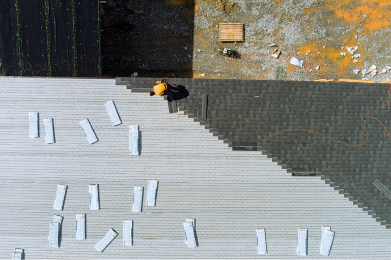 Aerial view of a person installing dark shingles on a roof, alongside a section of lighter material and scattered bundles.