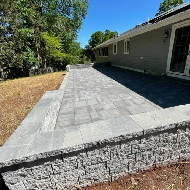 Newly constructed gray paver patio with retaining wall, next to a house with light green siding, under a blue sky.